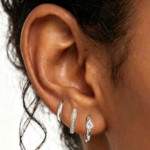 May include: Three silver hoop earrings are shown on an ear. The first is a plain, polished hoop. The second is a smaller hoop with a row of small, clear stones. The third is a small hoop with a single, clear stone.