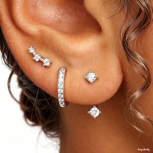 May include: A close-up of an ear adorned with multiple pieces of jewelry. There is a silver ear climber with three clear stones, a small hoop earring with clear stones, and two square-shaped stud earrings with clear stones. The jewelry is set against the backdrop of the ear.