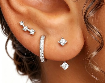 May include: A close-up of an ear adorned with multiple pieces of jewelry. There is a silver ear climber with three clear stones, a small hoop earring with clear stones, and two square-shaped stud earrings with clear stones. The jewelry is set against the backdrop of the ear.