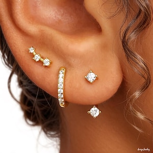 May include: A collection of gold earrings, including a curved bar with three clear stones, a small hoop with clear stones, and two square-cut stud earrings with clear stones. The earrings are displayed on an ear.