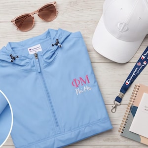 May include: A light blue Champion brand hooded windbreaker jacket with the Greek letters ΦM and the text Phi Mu embroidered in pink and white. Also included are a white baseball cap with a Φ logo, brown sunglasses, a blue Champion lanyard, and a notebook.