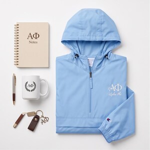 May include: A light blue hooded windbreaker with a half-zip closure and Alpha Phi embroidery, a beige spiral-bound notebook labeled AΦ Notes, a white mug with a laurel wreath AΦ logo, a metallic pen, and a brown leather keychain with an AΦ charm.