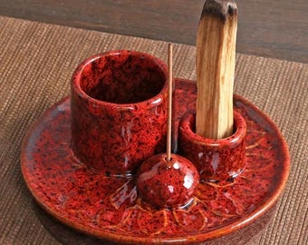 May include: A red ceramic tray with a speckled finish featuring a tall cylindrical holder, a smaller holder containing a piece of light brown wood, and a small spherical incense stick holder with a thin wooden incense stick inserted upright.