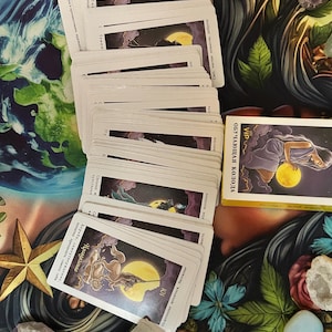 Könnte beinhalten: Ein Tarotkartenspiel mit Illustrationen von Figuren und Himmelskörpern. Die Karten sind fächerförmig angeordnet, mit einer Kartenbox auf der rechten Seite. Der Hintergrund umfasst Kristalle und ein florales Design.