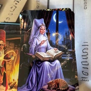 Könnte beinhalten: Eine rechteckige Tarotkarte mit der Aufschrift II. THE HIGH PRIESTESS, die eine Person in einem lavendelfarbenen Gewand und Kopfschmuck zeigt, die einen leuchtenden Zauberstab über ein offenes Buch hält, mit einer schlafenden Katze.