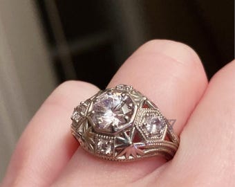May include: Close-up of a silver ring with a large, clear, round-cut center stone. The ring features intricate filigree and star-shaped designs, with smaller accent stones. The ring is set on a finger.