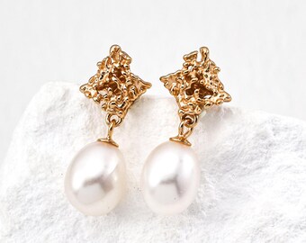 May include: A pair of gold-colored drop earrings. Each earring features a textured, square-shaped top with a dangling, oval-shaped white pearl. The earrings are displayed on a white surface.