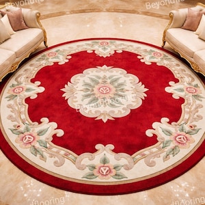 Könnte beinhalten: Ein runder roter Teppich mit einem verzierten Blumendesign. Der Teppich hat ein zentrales Medaillon und einen cremefarbenen Rand mit floralen Akzenten. Zwei cremefarbene Sofas mit goldenen Verzierungen befinden sich auf beiden Seiten des Teppichs.