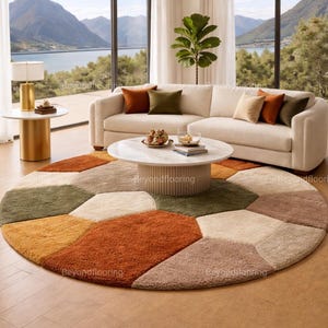 Könnte beinhalten: Ein modernes Wohnzimmer mit cremefarbenem Sofa, rundem Couchtisch und einem mehrfarbigen geometrischen Teppich. Der Teppich weist Farbtöne von Orange, Grün und Beige auf. Große Fenster bieten einen malerischen Blick auf einen See und Berge.