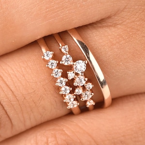 May include: A rose gold ring set consisting of two bands. One band features a cluster of small, round, clear stones arranged in a branching pattern, while the second band is a plain, polished rose gold metal band with a smooth, rounded surface.