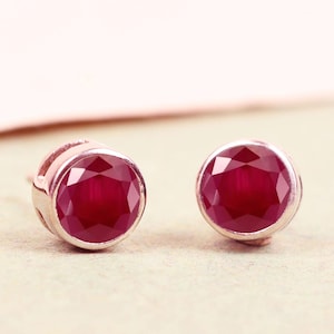 May include: A pair of stud earrings with round, faceted, deep red gemstones set in silver-coloured metal. The earrings are displayed on a light beige surface with a soft pink backdrop.