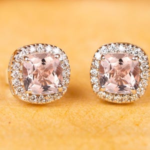 Puede incluir: Un par de pendientes de botón con una piedra preciosa de color rosa claro en talla cojín, rodeada por un halo de pequeñas piedras blancas redondas y transparentes engastadas en un marco metálico, creando un aspecto clásico.