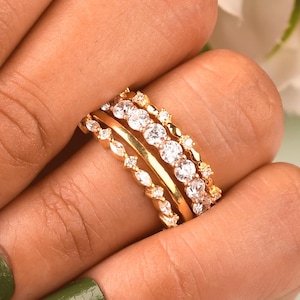 Puede incluir: Un conjunto de tres anillos dorados en un dedo. El set incluye una banda fina con un patrón texturizado en forma de marquesa, una banda lisa pulida y una banda con una hilera de piedras redondas transparentes en un diseño de metal festoneado.