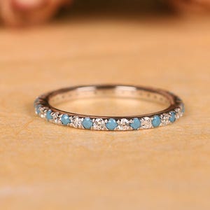 Op de afbeelding: Een zilveren ring met een band van kleine, ronde, lichtblauwe edelstenen en heldere stenen. De ring is geplaatst tegen een warme, neutrale achtergrond, met zachte, vage bloemelementen op de achtergrond.