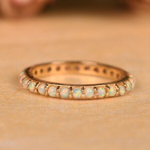 May include: A gold-colored ring with a band of small, round, white opals. The ring is set on a light brown surface, with blurred pink flowers in the background. The ring is a delicate piece of jewelry.