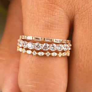 Puede incluir: Un conjunto de tres anillos dorados en un dedo. El anillo superior presenta una hilera de piedras en talla baguette, el anillo central tiene una serie de piedras en talla brillante redonda y el anillo inferior muestra un patrón de piedras en talla marquesa.