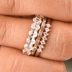Puede incluir: Un conjunto de tres anillos de oro rosa usados juntos. El primer anillo presenta una hilera de piedras claras de talla ovalada, el segundo es una banda con piedras rectangulares y el tercero es una banda delicada con pequeñas piedras claras redondas.