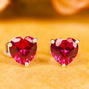 May include: A pair of heart-shaped stud earrings with faceted, deep red gemstones. The earrings are set in silver-colored metal and rest on a yellow-orange surface, with blurred flowers in the background.
