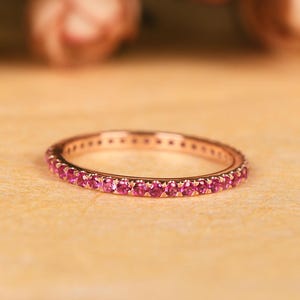 Pode incluir: Anel de ouro rosa com uma faixa de pequenas e vibrantes pedras preciosas rosa. O anel está sobre uma superfície bege clara, com flores rosa desfocadas ao fundo. Uma joia delicada.