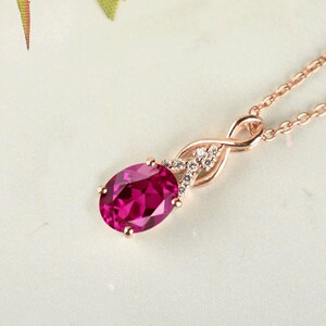 Könnte beinhalten: Eine Halskette aus Roségold mit einem oval geschliffenen, leuchtend pinkfarbenen Edelsteinanhänger. Die Fassung enthält eine zarte, unendlich geformte Schlaufe, die mit kleinen klaren Steinen besetzt ist und an einer dünnen Roségoldkette hängt.