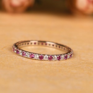 May include: A delicate ring with a band of alternating pink and clear gemstones. The ring is set on a light brown surface, with blurred pink flowers in the background. The ring is a piece of jewellery.