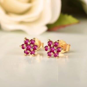 Peut inclure: Une paire de boucles d'oreilles à clous en or, chacune présentant un motif floral composé de six pierres précieuses rondes d'un rouge rosé éclatant. Les pierres sont serties dans une monture en or, formant un design floral délicat.