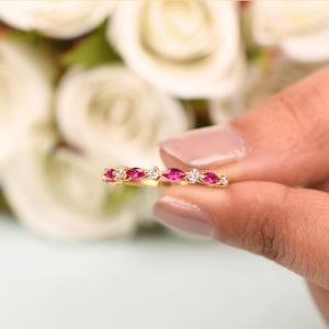May include: A gold ring featuring alternating marquise-cut pink gemstones and small round white gemstones. The ring is held in a person's hand, with a blurred background of white flowers.