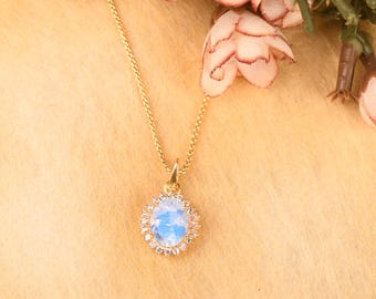 May include: A gold-colored necklace with a teardrop-shaped pendant. The pendant features a light blue stone surrounded by small, clear stones. The necklace is displayed on a light brown surface with pink flowers in the background.