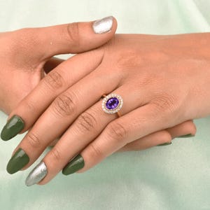 May include: A gold ring featuring an oval, deep purple gemstone surrounded by a halo of smaller, clear stones. The ring is set on a woman's hand with green and silver nail polish.