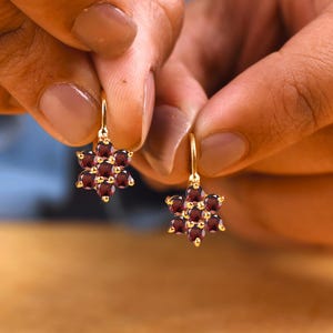 Pode incluir: Um par de brincos de ouro com um design floral composto por pedras preciosas granada vermelho-escuras. Cada brinco apresenta uma pedra central rodeada por seis pedras menores, suspensas por um gancho de ouro simples.