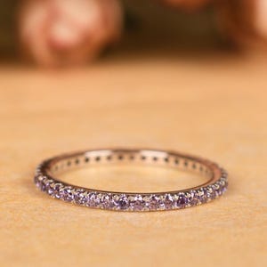May include: A delicate silver ring with a continuous row of small, light purple gemstones. The ring is set against a soft, neutral background, highlighting the sparkle of the gemstones. This elegant piece of jewelry is perfect for everyday wear or special occasions.