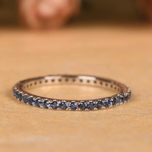 May include: A silver ring featuring a continuous row of small, round, blue gemstones. The ring is displayed against a soft, neutral backdrop, accentuating the sparkle of the gems. The ring is a full eternity band.