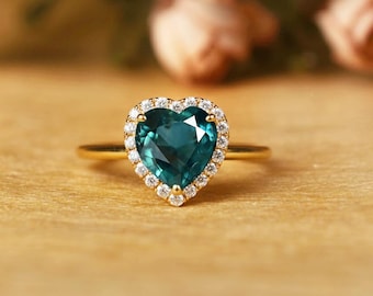 May include: A gold ring featuring a heart-shaped, teal gemstone surrounded by a halo of small, clear stones. The ring's band is a simple, polished gold. The setting and the band are both gold in color.