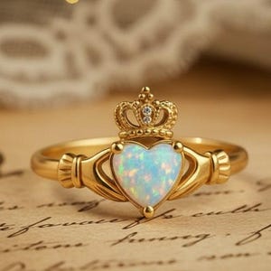 May include: A gold Claddagh ring with a heart-shaped opal centre stone. The ring features a crown and hands design, with detailed craftsmanship. The opal has a blue and white iridescent appearance. The ring is set on a vintage paper background.