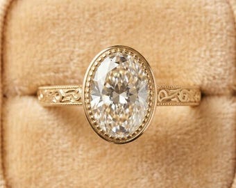May include: A gold engagement ring with an oval-cut diamond set in a bezel setting. The band features intricate filigree detailing. The ring is displayed in a beige velvet ring box, showcasing its elegant design.