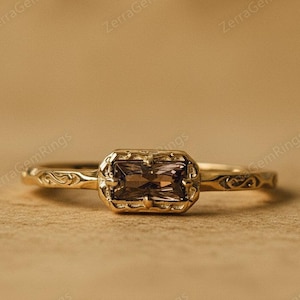 May include: A gold ring featuring a rectangular, brown gemstone. The ring has an intricate, filigree pattern around the stone and on the band. The background is a warm, neutral colour, accentuating the ring's details. This is a piece of jewellery.