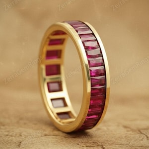 Könnte beinhalten: Ein goldener Bandring mit einer durchgehenden Reihe rechteckiger, tiefroter Edelsteine. Der Ring ist vor einem neutralen, strukturierten Hintergrund platziert. Der Ring ist im Full-Eternity-Stil gehalten.