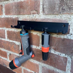 Może przedstawiać: Czarny uchwyt ścienny na wąż ogrodowy z dwoma dołączonymi dyszami. Uchwyt jest zamontowany na czerwonej ceglanej ścianie. Dysze są szare i czarne z pomarańczowymi akcentami.