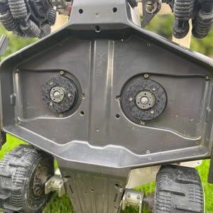 Puede incluir: Vista de cerca de un robot blanco y negro con múltiples ruedas. El robot tiene un diseño complejo con varios componentes mecánicos, incluyendo engranajes circulares y ruedas de oruga. El robot está posicionado sobre una superficie cubierta de hierba.