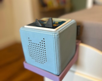 Puede incluir: Un reproductor de audio para niños en forma de cubo azul claro, con una rejilla de altavoz con forma de gato. El reproductor se encuentra sobre una base morada sujeta a una superficie blanca. Dos figuras grises con forma de tiburón están en la parte superior.