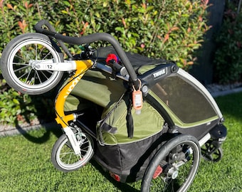 Può includere: Un passeggino da jogging Thule verde e nero con tre ruote, un telaio giallo e un manubrio nero. Il passeggino ha una copertura in rete ed è su un prato.