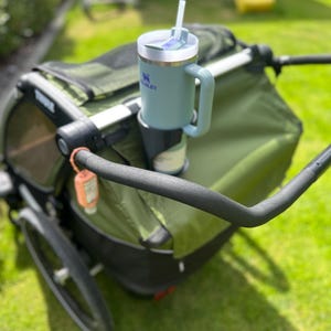 Könnte beinhalten: Ein hellgrüner Stanley-Becher mit Henkel und weißem Deckel, befestigt in einem Kinderwagen-Becherhalter. Der Kinderwagen ist olivgrün und schwarz, mit einem schwarzen Lenker. Der Hintergrund ist eine grüne Wiese und eine gelbe Rutsche.