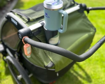 Può includere: Una tazza Stanley verde chiaro con manico e coperchio bianco, fissata in un portabicchieri per passeggino. Il passeggino è verde oliva e nero, con un manubrio nero. Lo sfondo è un prato verde e uno scivolo giallo.