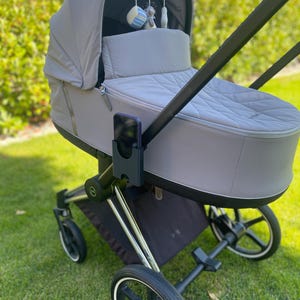 Può includere: Un passeggino grigio con telaio nero e dettagli argentati è posizionato su erba verde. Il passeggino ha un interno trapuntato e una capottina. Un mobile con giocattoli appesi è attaccato alla capottina.