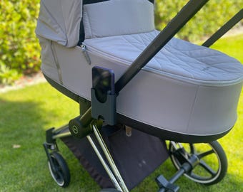 May include: A grey pram with a black frame and silver detailing is positioned on green grass. The pram features a quilted interior and a canopy. A mobile with hanging toys is attached to the canopy.