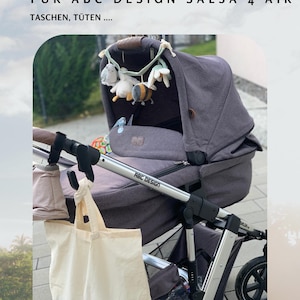 Op de afbeelding: Een donkergrijze kinderwagen met een crèmekleurige draagtas die aan de handgreep hangt. De kinderwagen heeft een bijpassende donkergrijze kap en een decoratieve speelgoedstang met pluche speelgoed. De tekst "HAKEN (1 STÜCK)" is zichtbaar.
