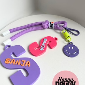 Può includere: Una collezione di portachiavi e accessori personalizzati. Include un cordino viola, due portachiavi a forma di lettera "S" con il nome "SANJA" in arancione e un ciondolo a forma di faccina sorridente viola. I portachiavi sono in tonalità viola, rosa e arancione.