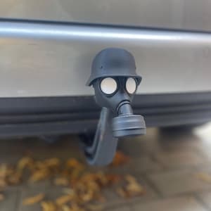 Op de afbeelding: Een zwarte trekhaakafdekking voor auto's met gasmasker en helm. De afdekking heeft de vorm van een gasmasker met een helm in Duitse stijl. Het masker heeft witte ooglenzen en een zwart ademhalingsapparaat.