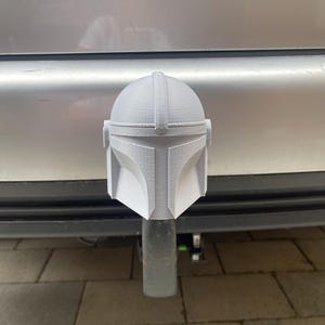 Op de afbeelding: Een grijze, 3D-geprinte Mandalorian helm trekhaakafdekking. De helm heeft het iconische T-vizier en de helmvorm, gemonteerd op een zwarte trekhaak. De afdekking is ontworpen voor een standaard 5 cm ontvanger.