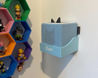 Puede incluir: Un altavoz azul claro con detalles negros en forma de orejas, montado en un soporte azul claro a juego. El nombre "Ilari" está impreso en el soporte. Una estantería hexagonal colorida con figuras es visible en el fondo.
